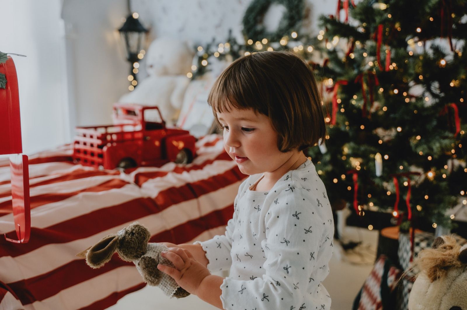 Bambina che gioca con un peluche sul letto a righe, davanti all'albero illuminato