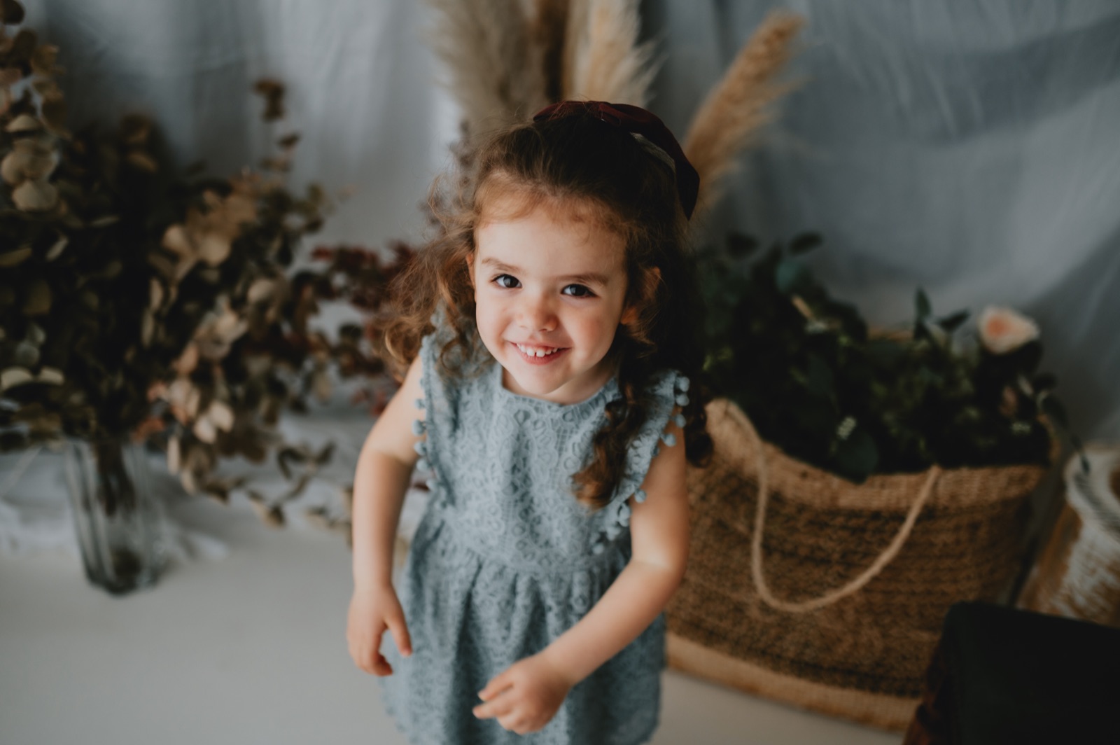 Bambina in tutina azzurra che sorride in piedi, fra i fiori secchi