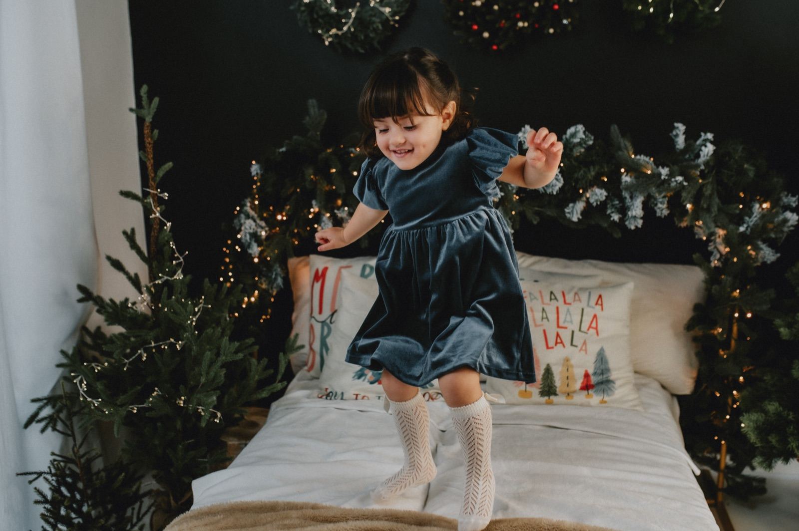 Bambina in abito di velluto blu che salta sul letto del set natalizio