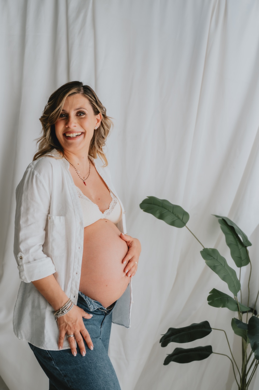 Futura mamma in camicia bianca e jeans che accarezza il pancione davanti a un fondale chiaro